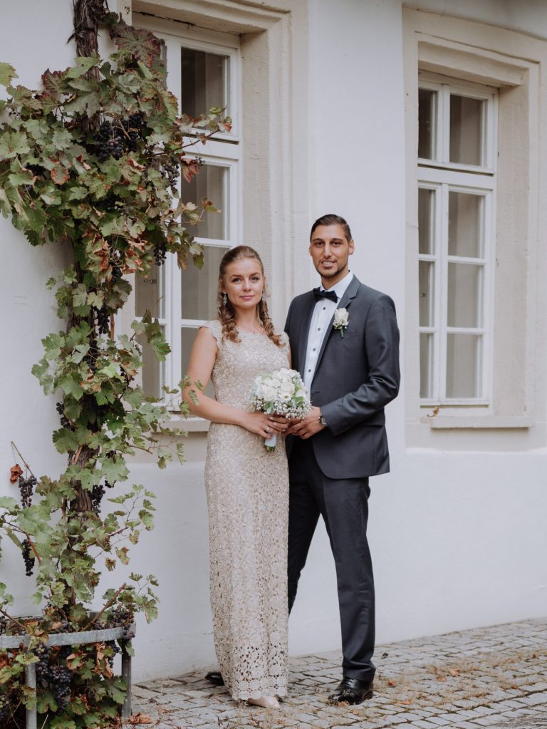 weddingvibesstudio, Brautpaar steht nebeneinander vor weißer wand mit Fenster und Weinreben